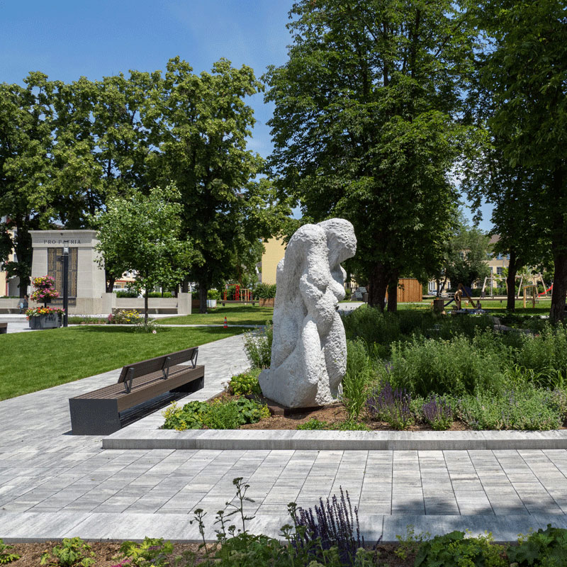 Mestský park Oberwart s kamennou sochou, lavičkou a zeleňou; Arret B25 VG4 kombinovaná dlažba, žulovo sivá tieňovaná, Friedl Steinwerke.