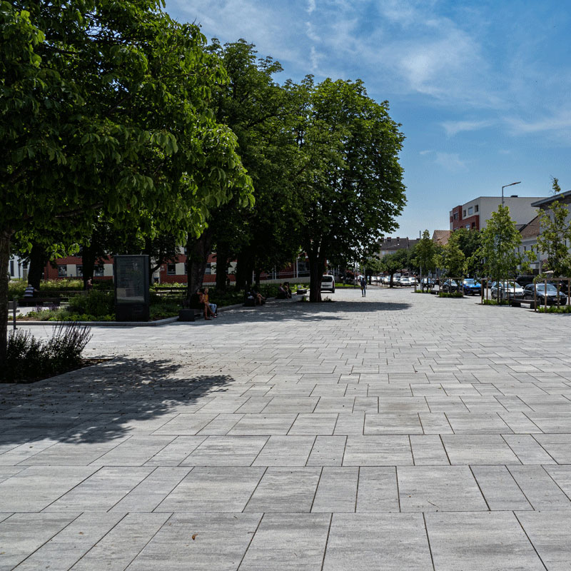 Mestský park Oberwart s rozsiahlym dláždeným priestorom a radom stromov; Magnum, žulovo sivá tieňovaná, Friedl Steinwerke.