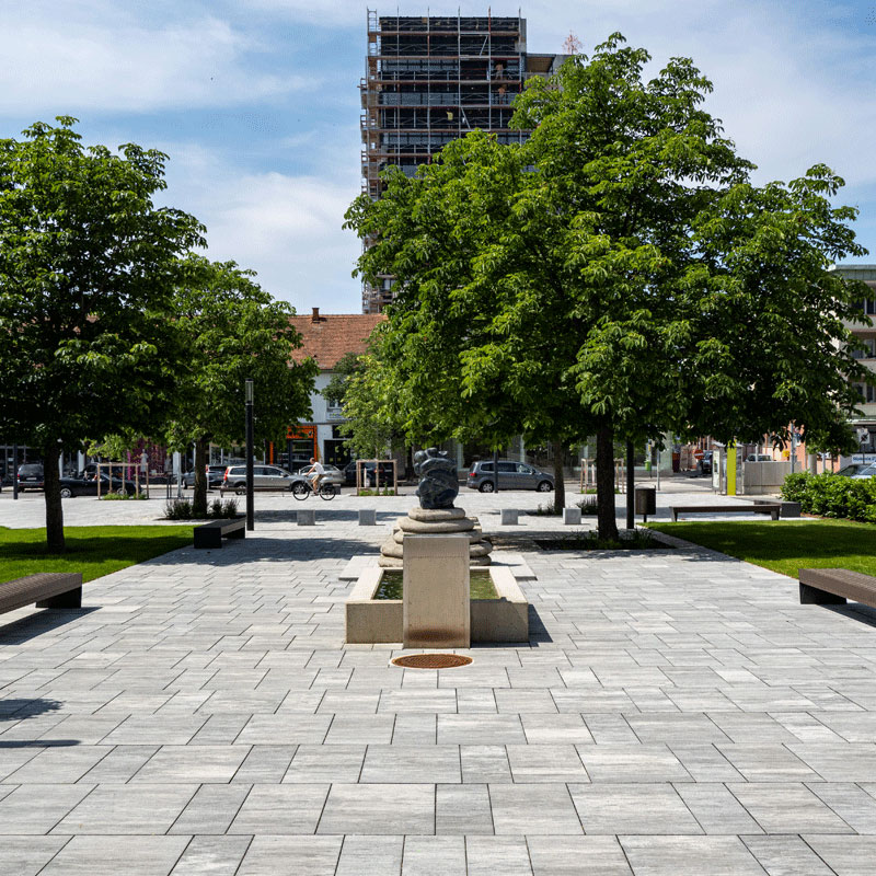 Mestský park Oberwart s fontánou a svetlou dláždenou plochou v tieni stromov; Magnum, žulovo sivá tieňovaná, Friedl Steinwerke.ontánou a dláždenou plochou v tieni stromov; Magnum a Arret B25 VG4 dlažba, žulovo sivá tieňovaná, Friedl Steinwerke.